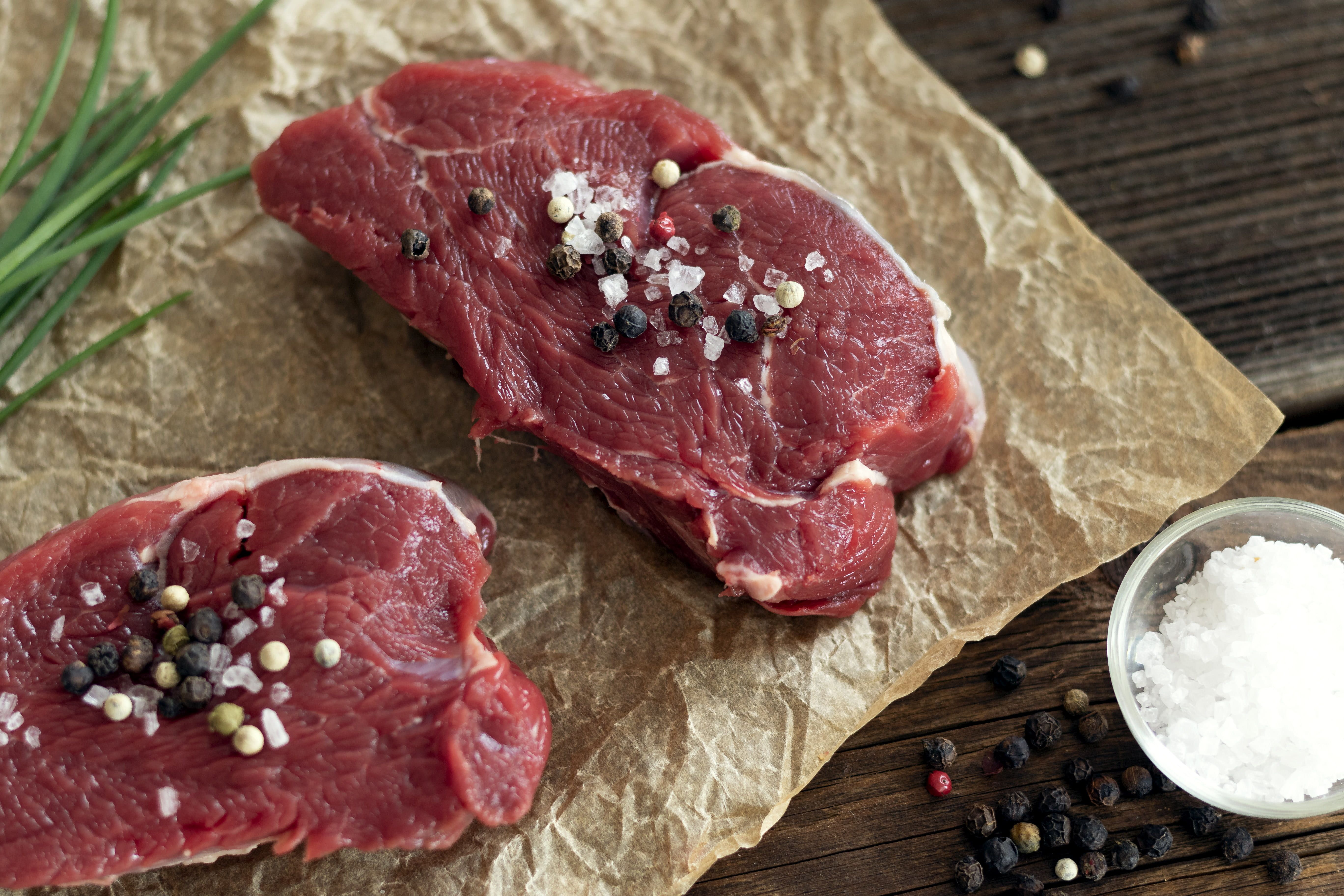 Zwei Stück rohes Rindfleisch mit Salz und Pfeffer bestreut auf Backpapier, daneben Schnittlauch, eine Schale Salz und Pfeffer.