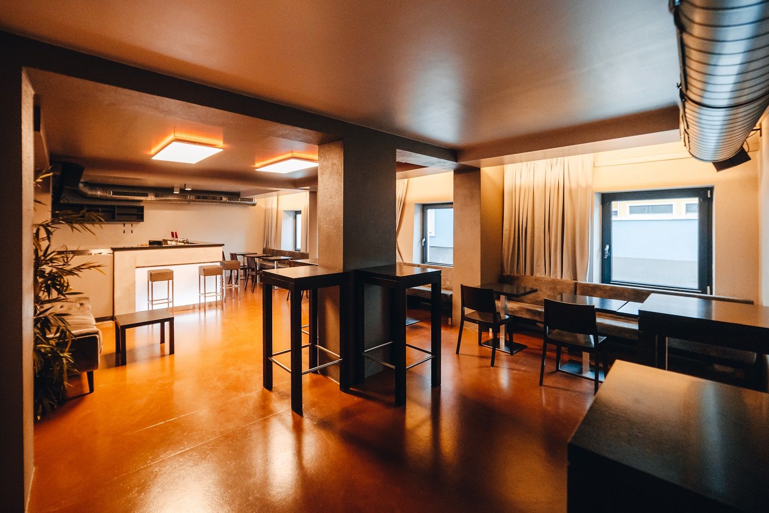 Modern lounge with bar, high tables and chairs, warm light and large windows.