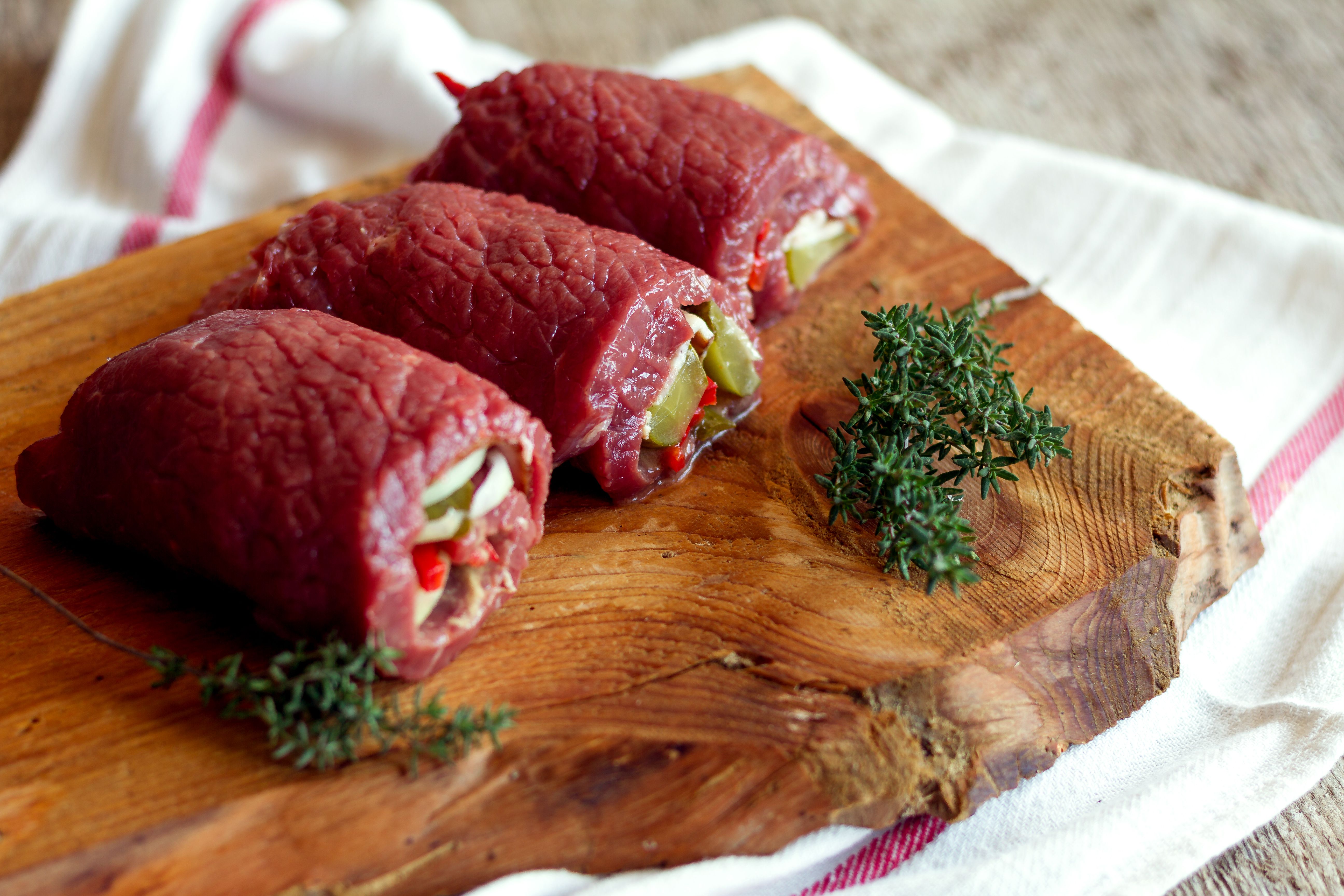 Drei gefüllte rohe Rinderrouladen auf einem Holzbrett mit Thymianzweigen.