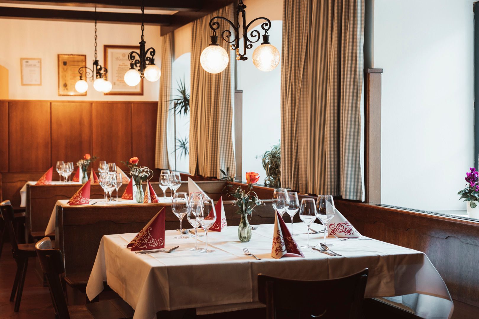 Eine gemütliche Gaststube mit gedeckten Tischen, Holzbänken, Lampen und Vorhängen.