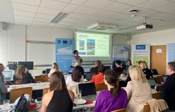 In einem Schulungsraum sitzen mehrere Teilnehmer eines Workshops mit dem Rücken zur Kamera in mehreren Reihen. Davor steht eine Person vor mehreren Roll-Ups und einer Leinwand.