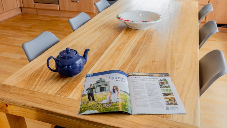 Ein Holztisch mit einer blauen Teekanne, einer Schüssel und einer Zeitschrift in einer Küche.