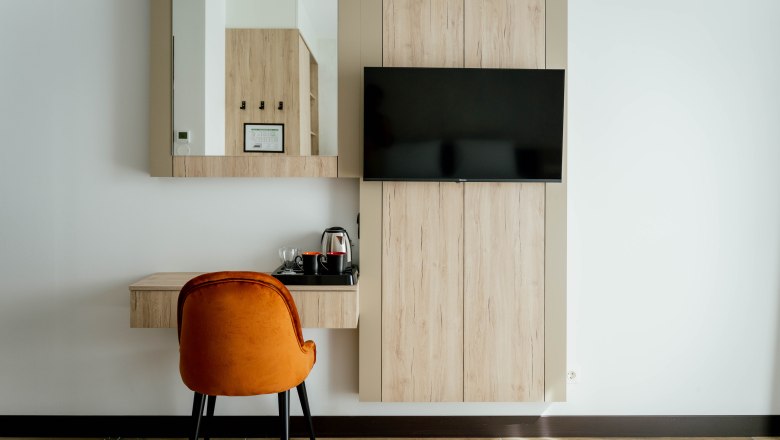 Modernes Hotelzimmer mit Holzmöbeln, einem Spiegel, einem Fernseher und einem orangefarbenen Stuhl.