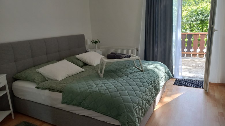 Ein gem&uuml;tliches Schlafzimmer mit einem grauen Bett, gr&uuml;ner Bettdecke und offenem Balkon mit Blick ins Gr&uuml;ne.