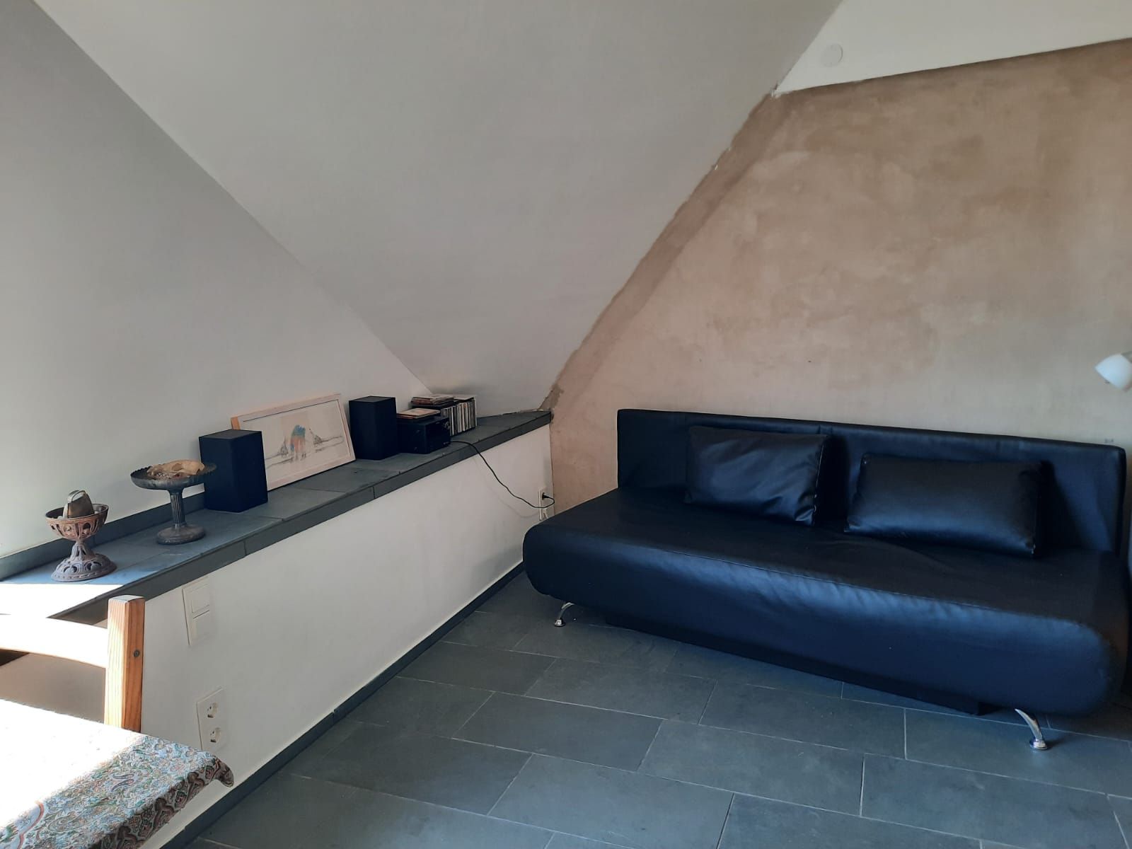 Ein schlicht eingerichtetes Zimmer mit einem schwarzen Sofa, einer Schräge und einem Regal mit Dekorationen.