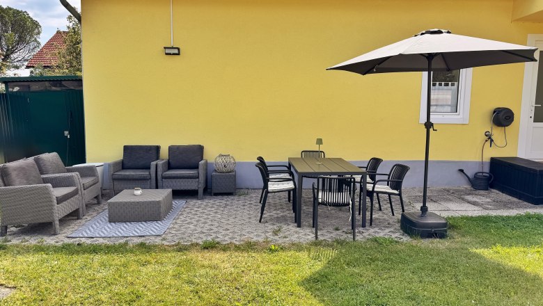 Terrasse mit gelber Hauswand, grauen Sitzmöbeln, Esstisch, Stühlen und Sonnenschirm.