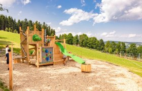 Spielplatz mit Klettergerüst und Rutsche auf einer Wiese, umgeben von Bäumen und blauem Himmel.