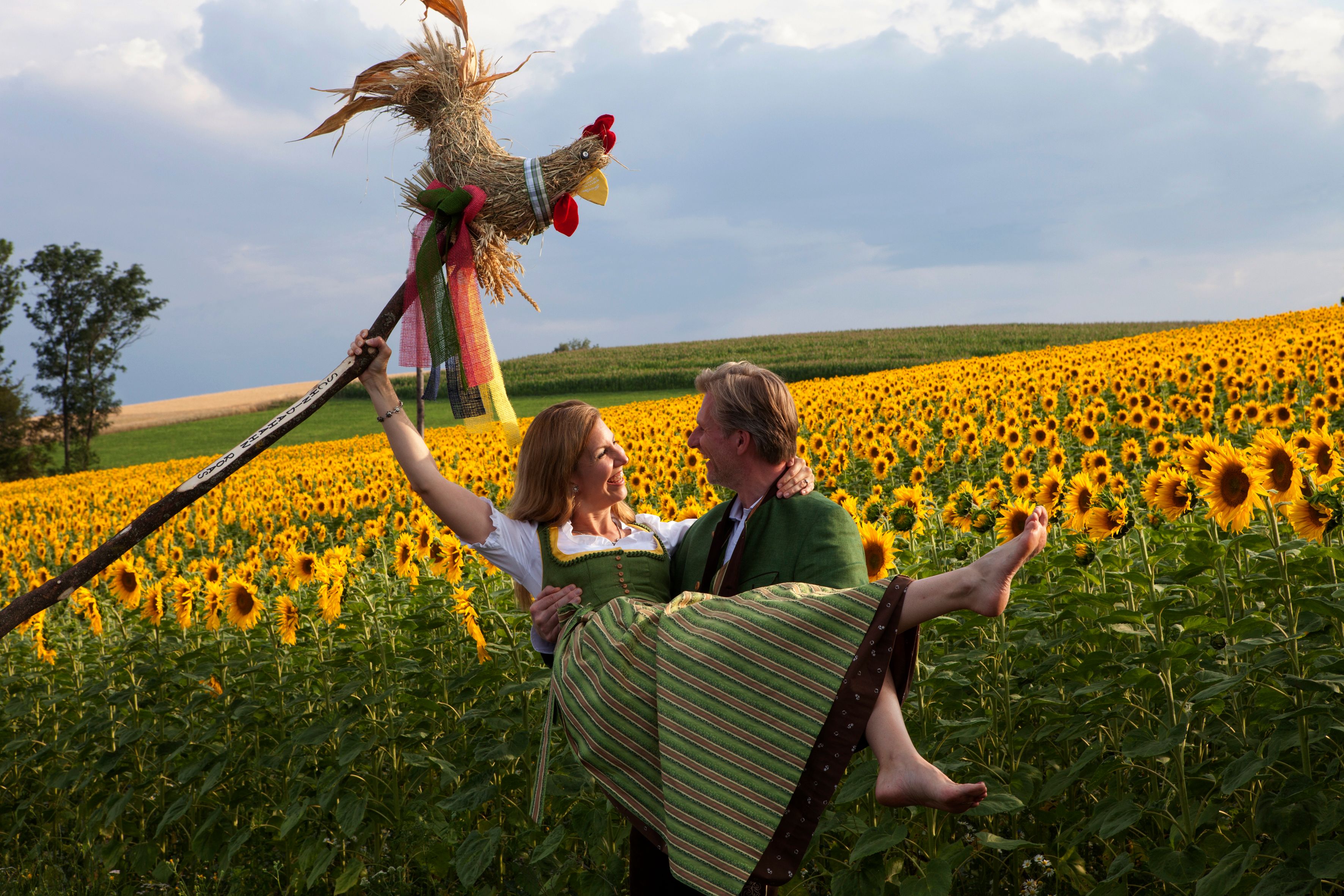 Mann trägt Frau im Arm, beide in Tracht, vor einem Sonnenblumenfeld.
