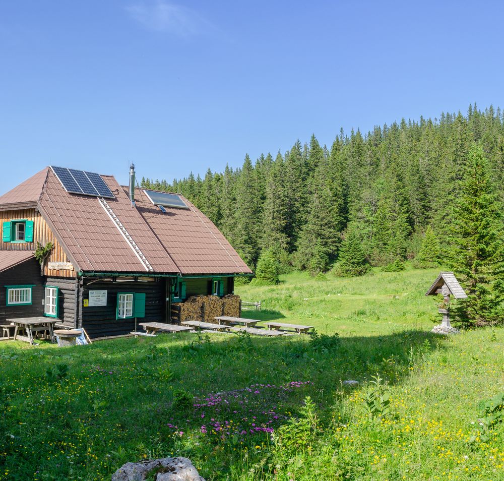 A mountain cabin with solar panels on the roof, surrounded by green meadows and woods.