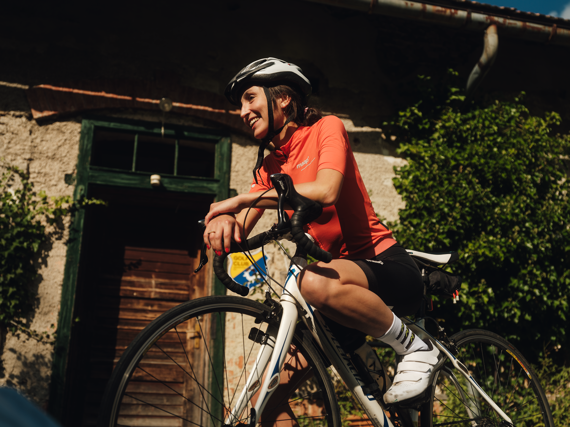 Ein strahlender Sommertag lädt zu einer erfrischenden Radtour ein. Die Radfahrerin genießt die Freiheit und die atemberaubende Landschaft, während sie an einem charmanten, rustikalen Gebäude vorbeifährt, umgeben von üppigem Grün.