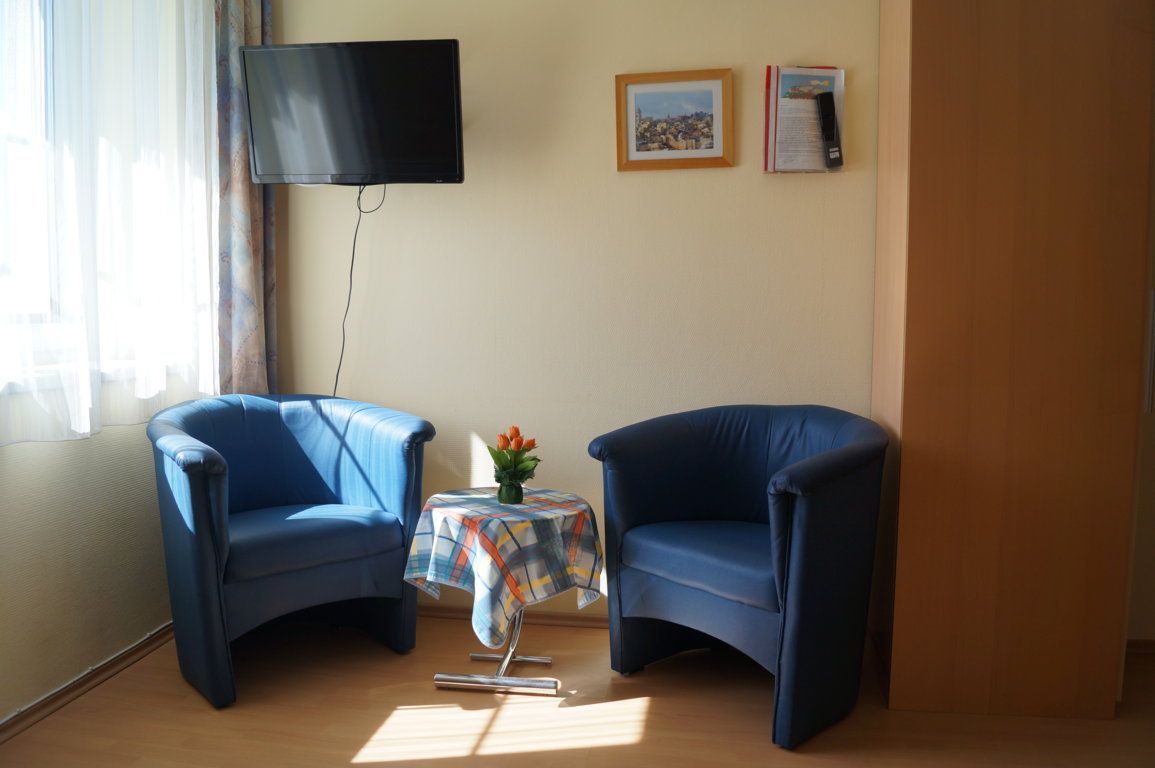 Zwei blaue Sessel mit einem kleinen Tisch dazwischen, darauf eine Vase mit Blumen. Ein Fernseher hängt an der Wand, daneben zwei Bilder.