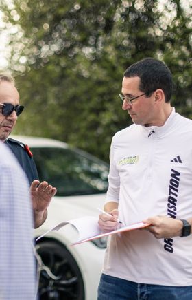 Ein Mann mit dunklen Haaren und weißen Sportshirt mit der Aufschrift Organisation hält ein Clipboard in der Hand und spricht mit einem Mann von der Feuerwehr.