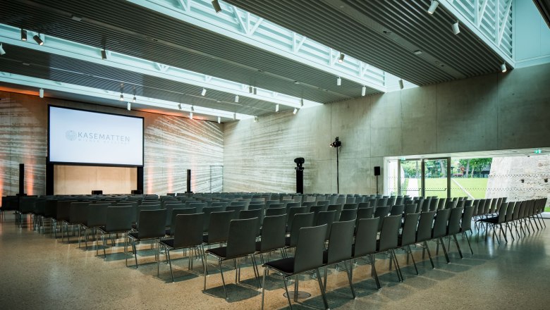 Moderner Veranstaltungsraum mit Stuhlreihen und gro&szlig;er Leinwand, beschriftet mit 'Kasematten Wiener Neustadt'.
