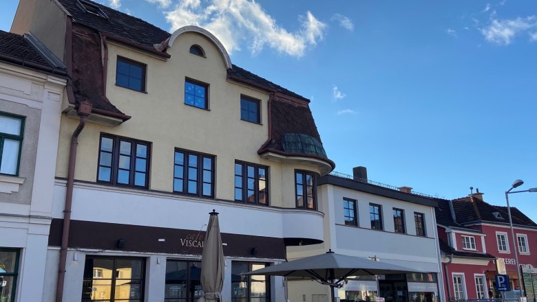 Außenansicht eines Cafés mit Terrasse und Sonnenschirm vor einem mehrstöckigen Gebäude.