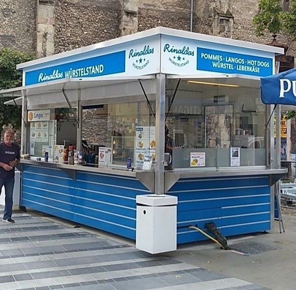 Ein blauer Imbissstand namens Rinaldas Würstelstand mit Angeboten wie Pommes und Hot Dogs.