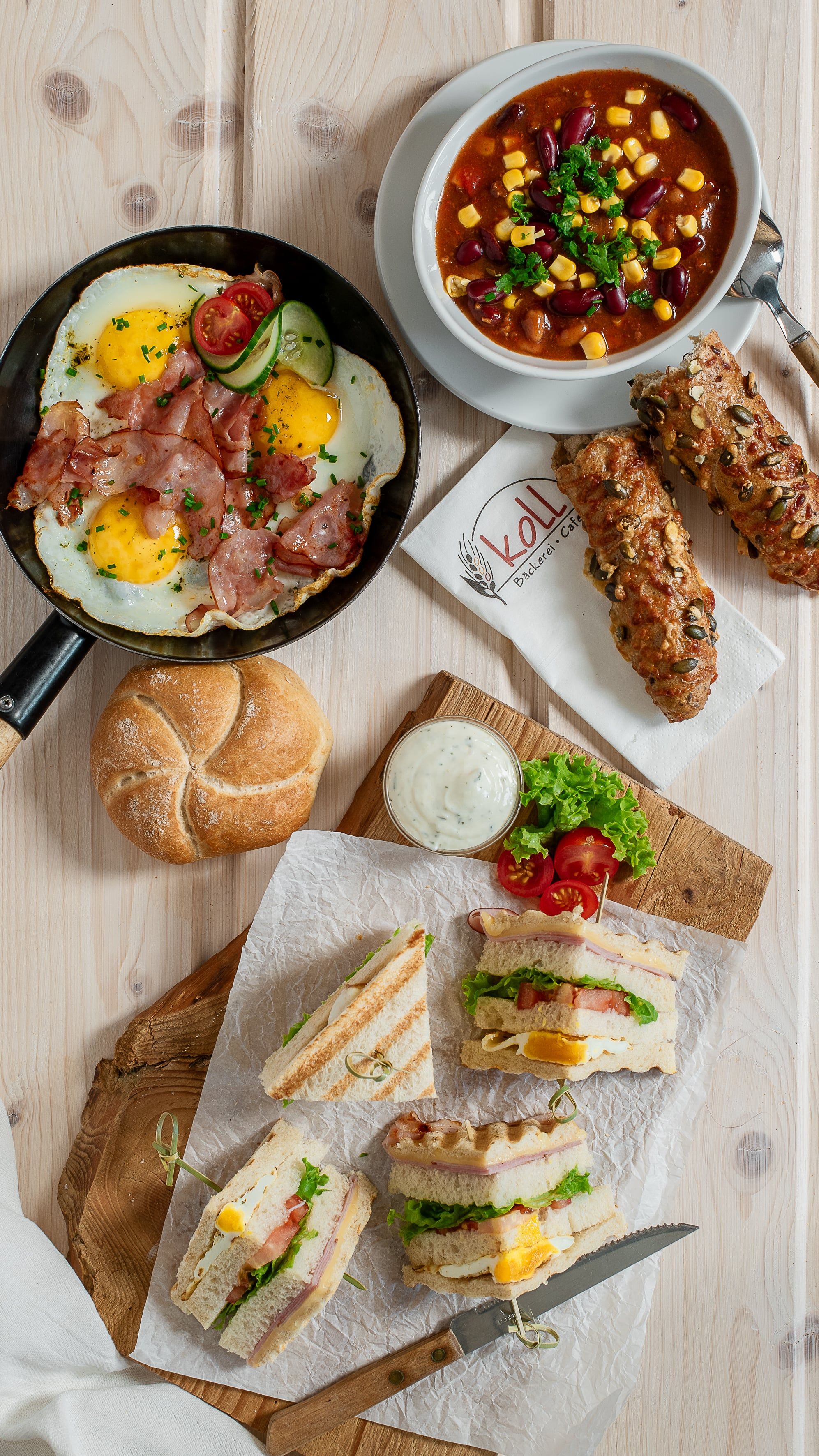 Verschiedene warme Snacks auf einem Tisch, darunter Sandwiches, Spiegelei mit Speck, Suppe und Brötchen.