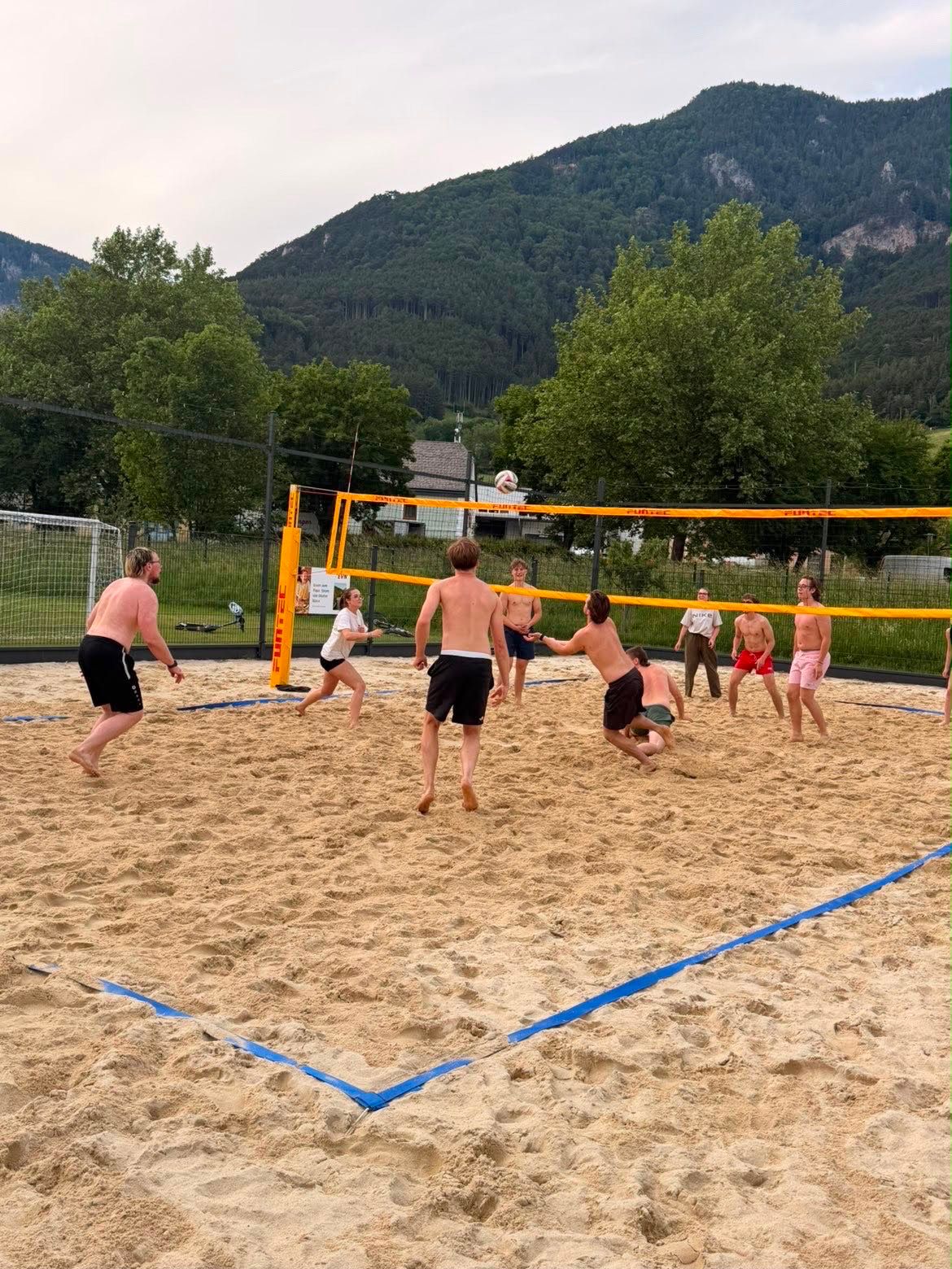 Group of people playing beach volleyball on a sand court with mountains in the background.