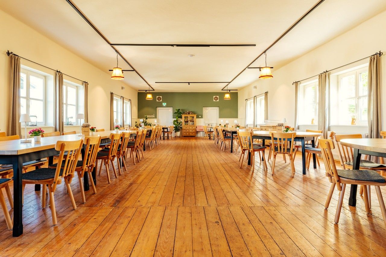 Ein langer Saal mit runden und eckigen Tischen, Stühlen, großen Fenstern mit Vorhängen und Holzboden.