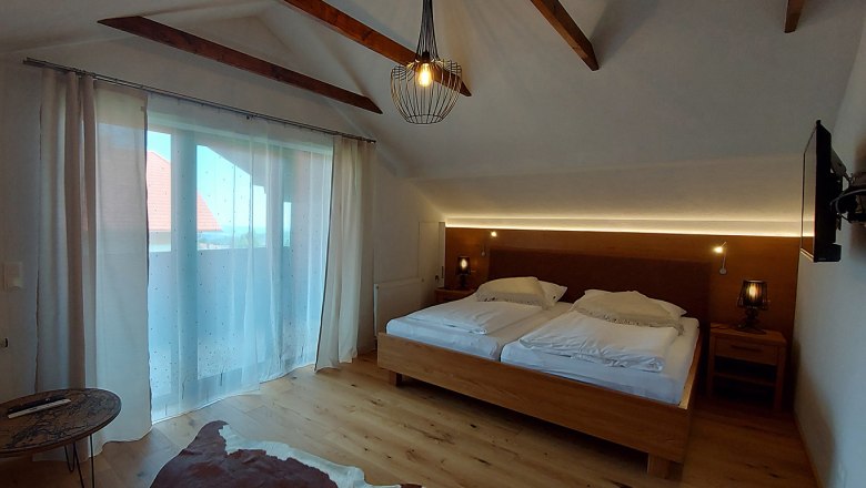 Ein gemütliches Doppelzimmer mit Holzboden, einem großen Bett, Vorhängen und einem kleinen Tisch. Eine Lampe hängt von der Decke.