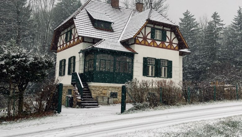 Orlet an der Rax, © Sofia Orlet Ein verschneites Haus im traditionellen Stil mit grünen Fensterläden, umgeben von Bäumen und einer schneebedeckten Straße.