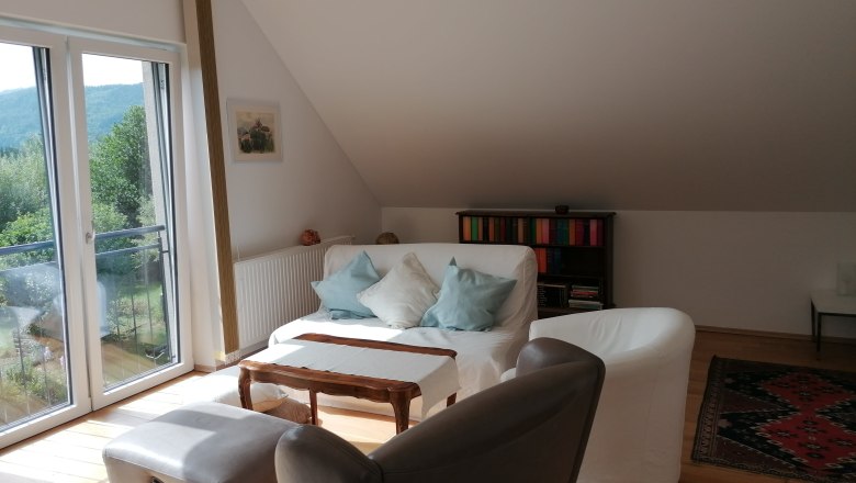 Heller Wohnbereich mit Sofa, Sesseln, Bücherregal und großem Fenster mit Blick ins Grüne.