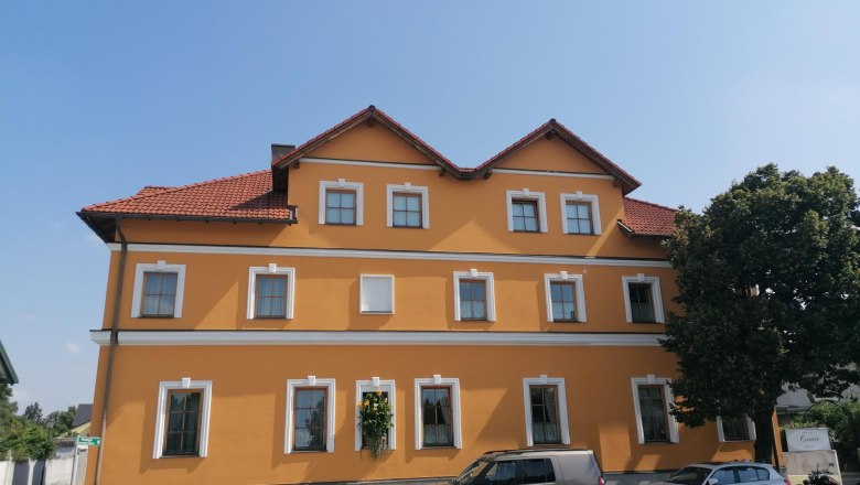 Gelbes Gebäude mit roten Dachziegeln und mehreren Fenstern, davor geparkte Autos und ein Baum.