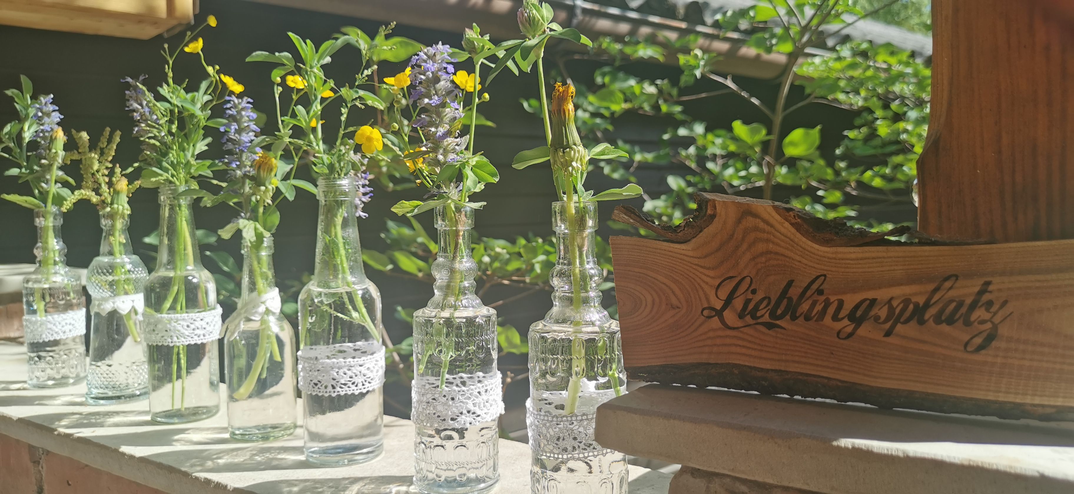 Kleine Glasflaschen mit Wiesenblumen und ein Holzschild mit der Aufschrift 'Lieblingsplatz'.