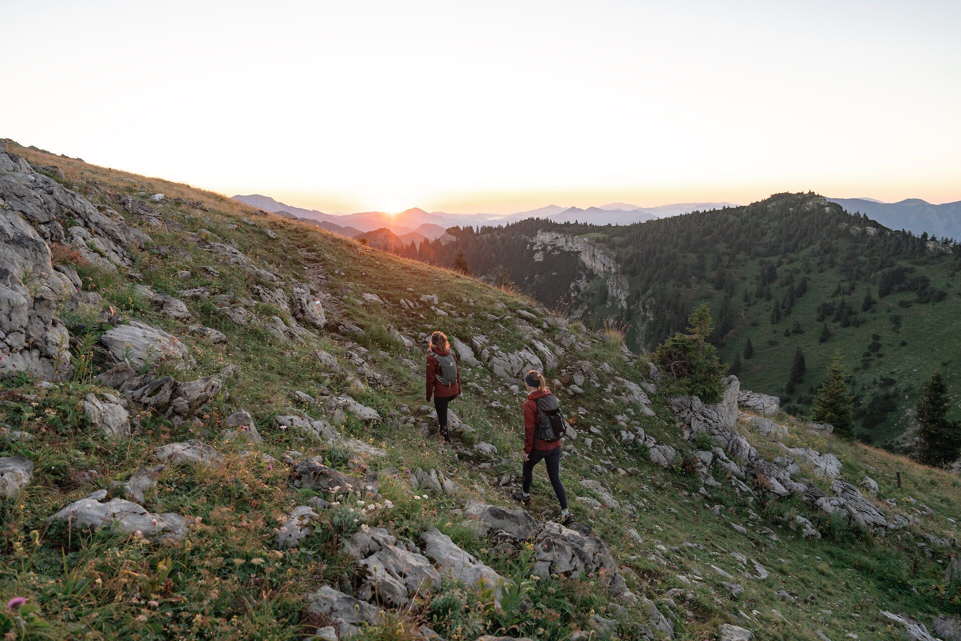 Die sanften Hügel der Ybbstaler Alpen erstrahlen im warmen Licht des Sonnenaufgangs, während zwei Wanderer den Pfad entlang schreiten. Die frische Bergluft und die atemberaubende Aussicht auf die umliegenden Gipfel schaffen eine unvergessliche Atmosphäre der Freiheit und des Abenteuers.