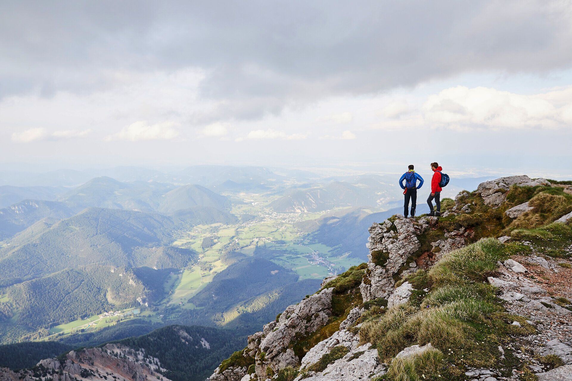 Die majestätischen Berge erstrecken sich bis zum Horizont, während zwei Wanderer auf dem Gipfel stehen und die atemberaubende Aussicht genießen. Die frische Bergluft und die sanften Wiesen laden dazu ein, die Schönheit der Natur in vollen Zügen zu erleben.