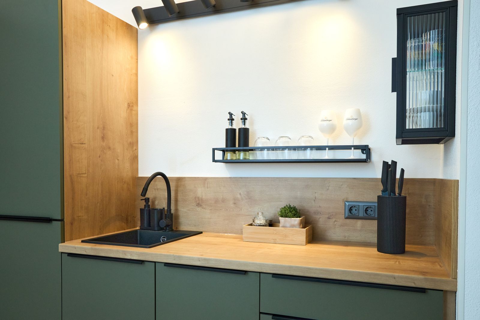 Modern, fully equipped kitchen unit with green fronts, wooden worktop, black sink and mixer tap as well as open shelves with glasses and kitchen utensils.