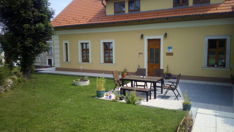 Ein gelbes Haus mit rotem Dach und Terrasse, umgeben von Rasen und Pflanzen.