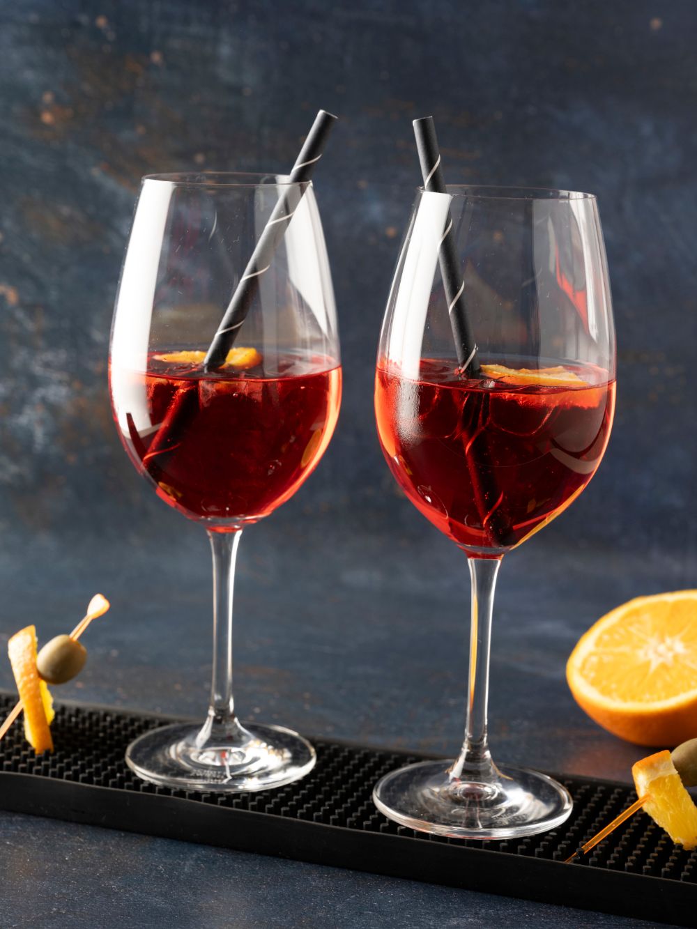 Zwei Weingläser mit Martini Vibrante Sprizz, Orangen und Strohhalmen, daneben Spieße mit Orangen und Oliven.