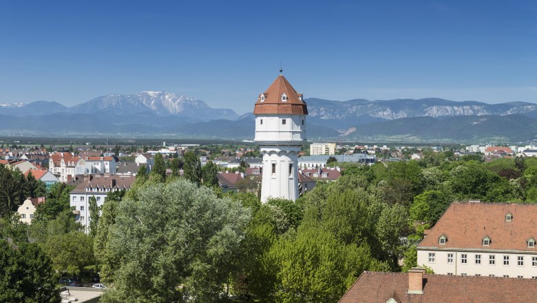 Ein wei&szlig;er Wasserturm mit rotem Dach in einer gr&uuml;nen Stadtlandschaft vor einem Bergpanorama.