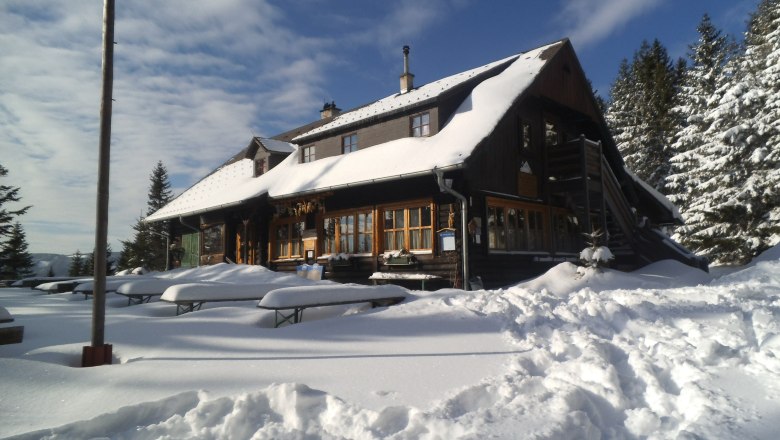 Verschneite Bergh&uuml;tte mit Holzb&auml;nken im Vordergrund und Tannen im Hintergrund.
