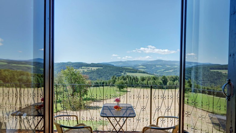 View from a balcony of a hilly landscape with forests and fields, two chairs and a table with flowers in the foreground.