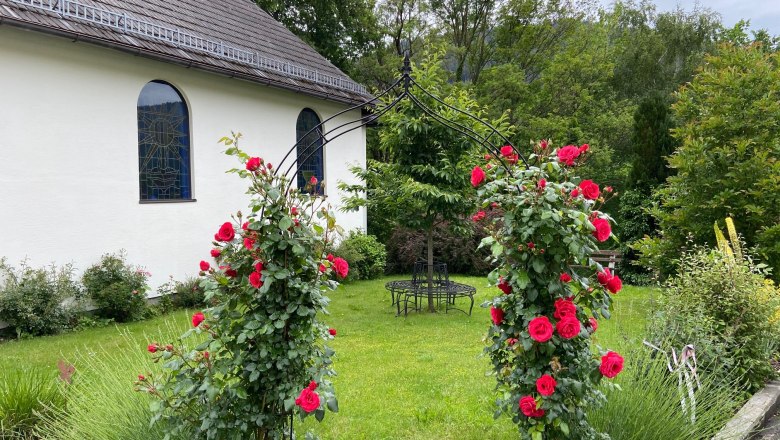 A rose arch with red roses in front of a chapel with colorful windows and a green garden.