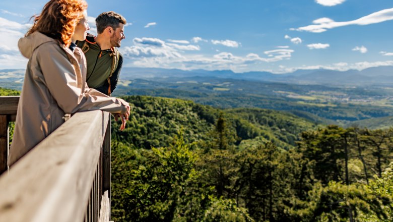 Zwei Personen stehen auf der Aussichtswarte und blicken &uuml;ber eine bewaldete, h&uuml;gelige Landschaft.