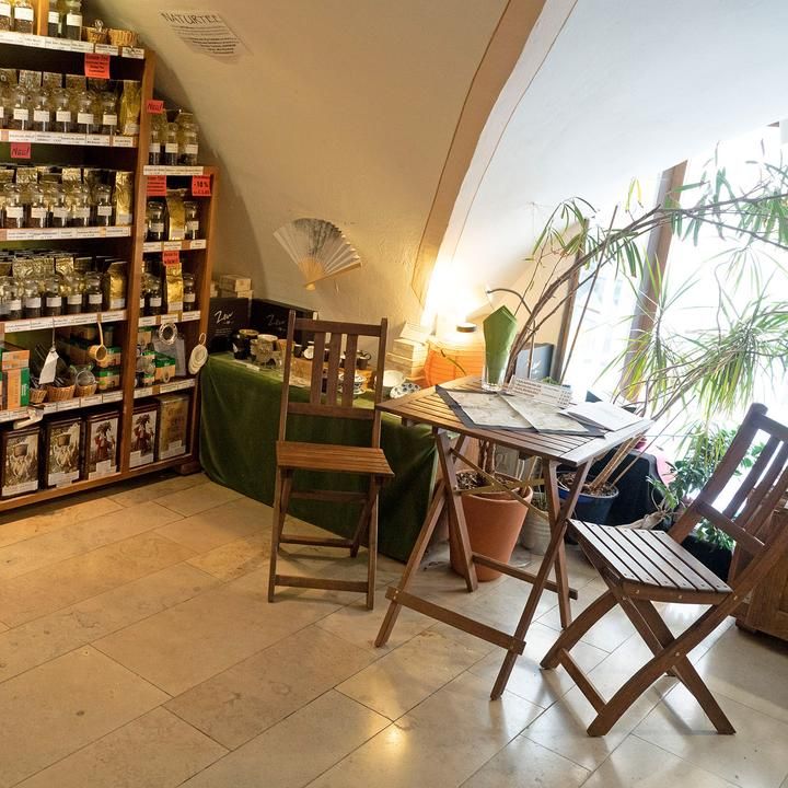 Ein gemütlicher Teeladen mit Holzregalen voller Teepackungen, einem kleinen Tisch und Stühlen sowie Pflanzen am Fenster.