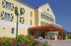 Außenansicht eines gelben Hotels mit blauen Fensterläden und einem Vordach. Vor dem Gebäude blühen gelbe und pinke Blumen.