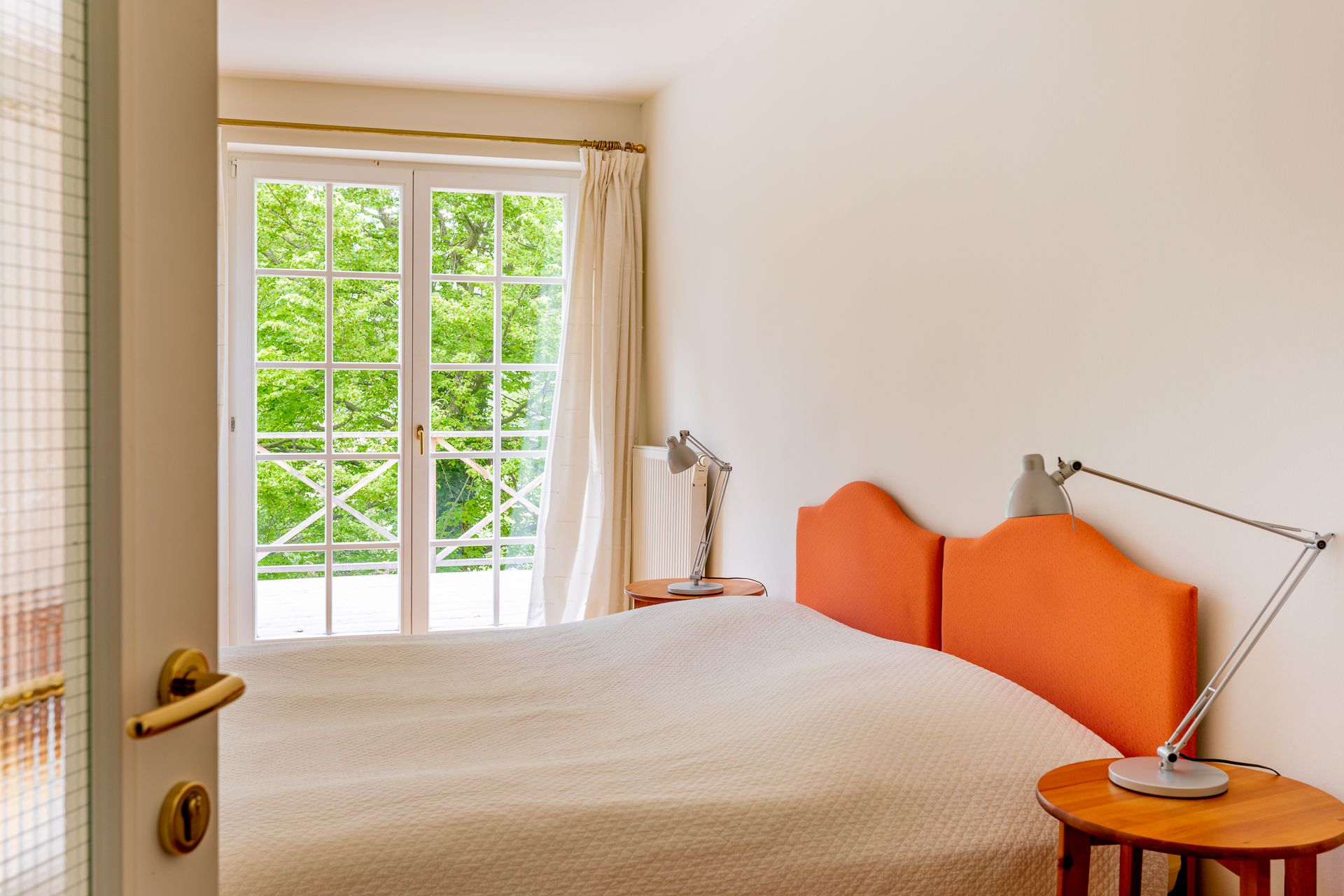 Ein Schlafzimmer mit einem orangefarbenen Kopfteil, einem Doppelbett und einem Fenster mit Blick auf grüne Bäume.