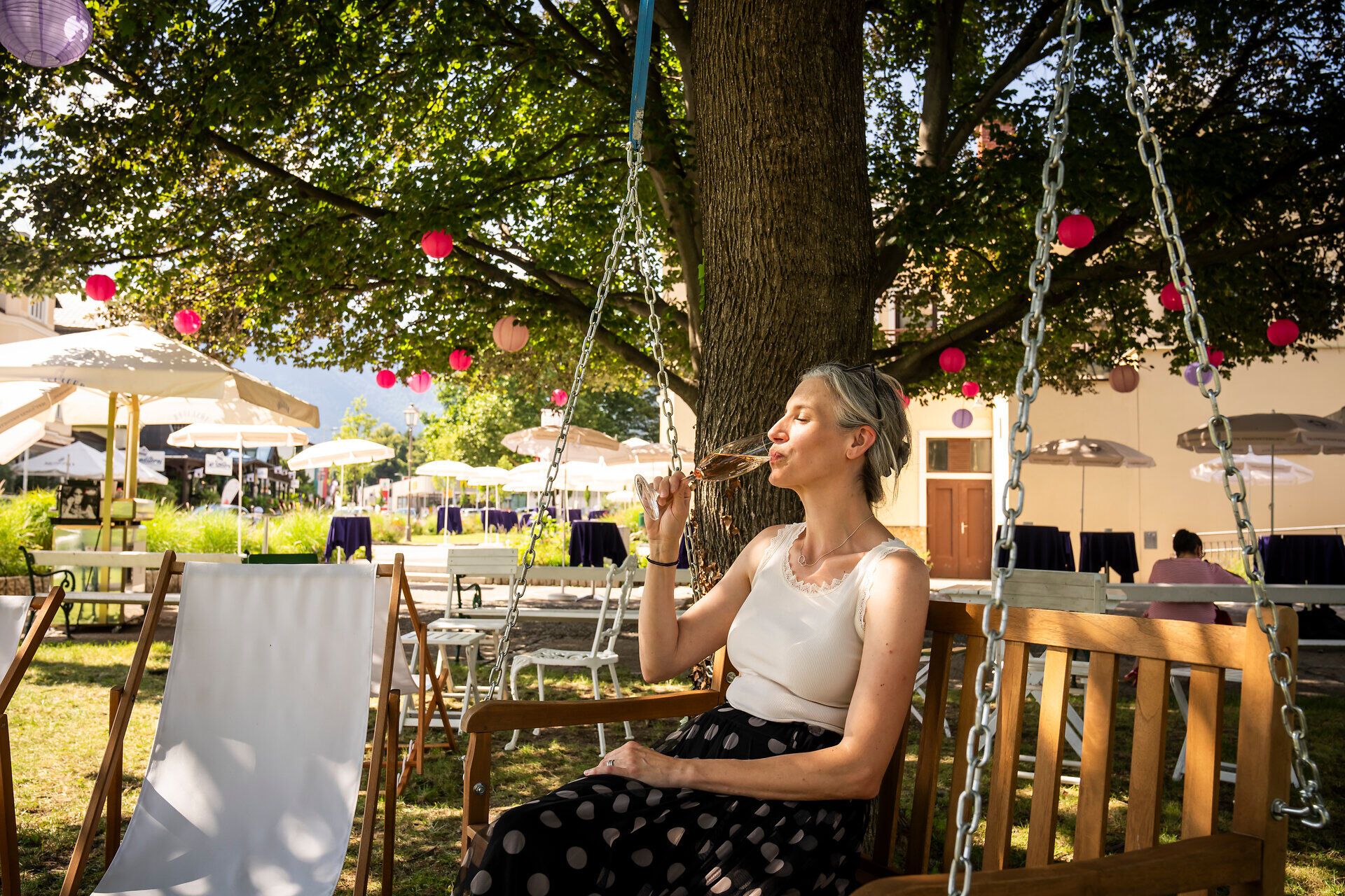 Unter dem schattenspendenden Baum genießen Besucher eine entspannte Auszeit mit einem Glas Sekt. Die sanfte Brise und die festliche Atmosphäre laden dazu ein, die Schönheit des Sommers in den Wiener Alpen zu erleben.