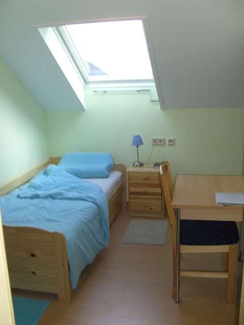 Ein kleines Einzelzimmer mit Bett, Nachttisch, Schreibtisch und Stuhl unter einem Dachfenster.