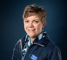 Portraitfoto Wiener Alpen Mitarbeiterin, die eine Jacke mit Wiener Alpen Logo trägt und vor einem dunklem Hintergrund steht.