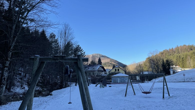 Snow-covered playground with swings, surrounded by trees and mountains under a clear blue sky.