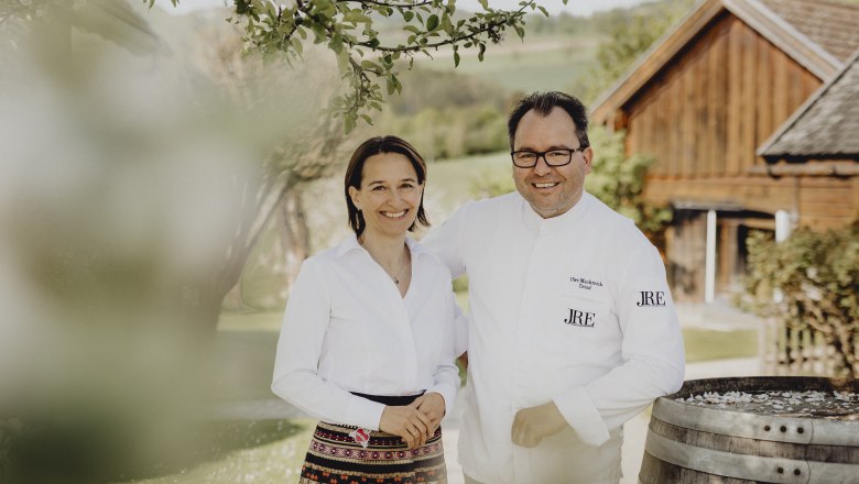 A man and a woman stand smiling outside in front of a wooden building.
