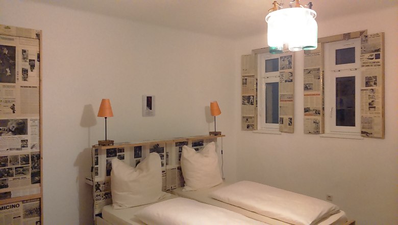 Rooms at Hotel Binder, © Wiener Alpen Hotel room with double bed, newspaper decorations on the walls and windows, two orange lamps.