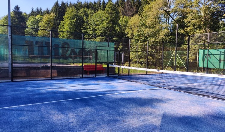 Padelplatz mit blauem Boden und Netz, umgeben von Bäumen.
