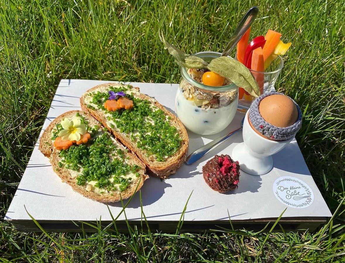 Frühstücksbrett im Gras mit zwei belegten Broten, Joghurt im Glas, Gemüsesticks, gekochtem Ei im Eierbecher und einer kleinen Süßigkeit.