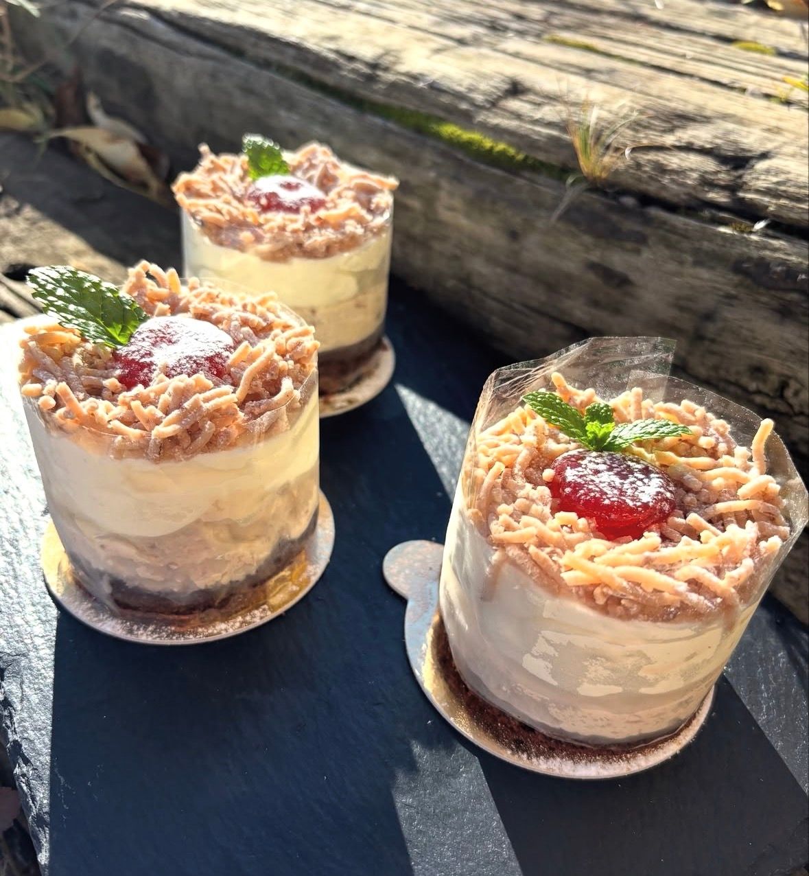 Drei kleine Desserttörtchen mit Creme, Krokant und roter Fruchtgarnitur auf dunklem Untergrund im Sonnenlicht.