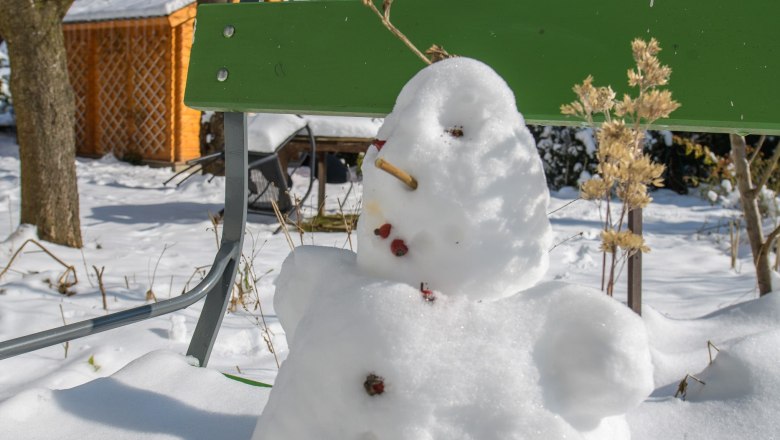 Ein kleiner Schneemann sitzt auf einer grünen Bank im verschneiten Garten.